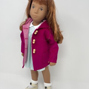 Pu&ograve; includere: Una bambola con lunghi capelli ramati indossa un cappotto magenta con cappuccio e alamari chiari, un vestito a righe bianche e azzurre con colletto bianco, calzini bianchi a coste e scarpe color crema con un bottone scuro.