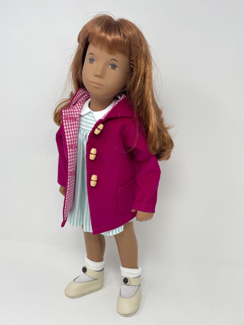 Pu&ograve; includere: Una bambola con lunghi capelli ramati indossa un cappotto magenta con cappuccio e alamari chiari, un vestito a righe bianche e azzurre con colletto bianco, calzini bianchi a coste e scarpe color crema con un bottone scuro.