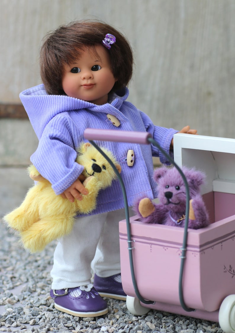 Pu&ograve; includere: Una bambola con capelli castani e una molletta viola, indossa una giacca con cappuccio in velluto a coste color lavanda con alamari, pantaloni bianchi e scarpe viola. La bambola tiene un orsetto giallo accanto a una carrozzina giocattolo rosa.