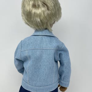 May include: A doll wearing a light blue denim jacket with white stitching and dark blue pants. The doll has short, light blonde hair. The back of the doll is facing the camera.