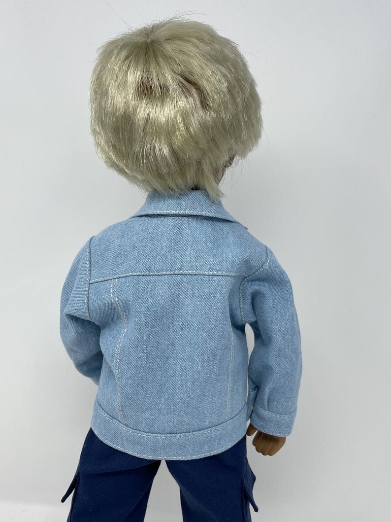 May include: A doll wearing a light blue denim jacket with white stitching and dark blue pants. The doll has short, light blonde hair. The back of the doll is facing the camera.