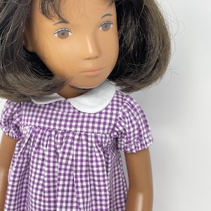Può includere: Una bambola con capelli castani e occhi marroni, che indossa un vestito a quadri viola e bianco con colletto bianco. La bambola ha un incarnato chiaro e guarda leggermente di lato.