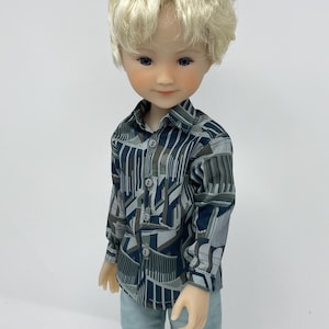May include: A doll with blonde hair, blue eyes, and a fair complexion. The doll wears a long-sleeved button-down shirt featuring a dark blue, grey, and brown geometric pattern, paired with light blue corduroy trousers with a vertical texture.