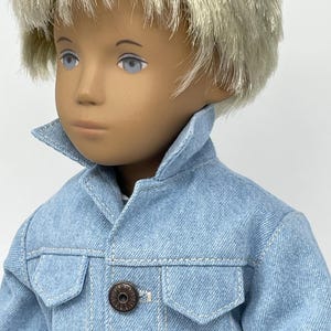 May include: A doll with light blonde hair and blue eyes wearing a light blue denim jacket with two front pockets and two bronze-colored buttons. The doll's skin tone is fair, and the jacket has visible stitching.