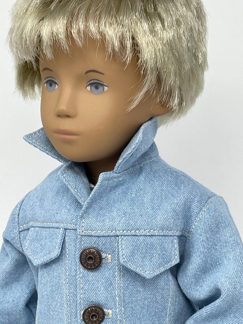 May include: A doll with light blonde hair and blue eyes wearing a light blue denim jacket with two front pockets and two bronze-colored buttons. The doll's skin tone is fair, and the jacket has visible stitching.