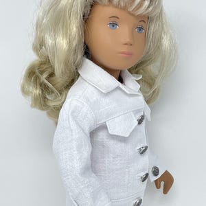 May include: A doll with blonde hair and blue eyes wearing a white jacket with silver buttons. The jacket has a textured fabric and a small pocket on the chest. The doll is wearing a matching white outfit.