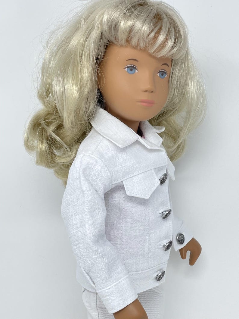 May include: A doll with blonde hair and blue eyes wearing a white jacket with silver buttons. The jacket has a textured fabric and a small pocket on the chest. The doll is wearing a matching white outfit.