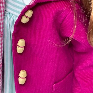 Pu&ograve; includere: Una giacca in velluto a coste color magenta brillante con tre bottoni a levetta in legno chiaro. Sotto, si vede un vestito a righe azzurre e bianche con colletto bianco. Un tessuto a quadri rosa e bianco &egrave; parzialmente visibile a sinistra.