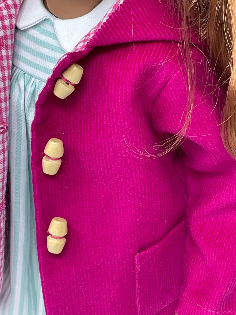 Pu&ograve; includere: Una giacca in velluto a coste color magenta brillante con tre bottoni a levetta in legno chiaro. Sotto, si vede un vestito a righe azzurre e bianche con colletto bianco. Un tessuto a quadri rosa e bianco &egrave; parzialmente visibile a sinistra.
