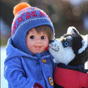 Pu&ograve; includere: Una bambola con capelli castani indossa un cappotto in velluto a coste blu, un berretto in maglia blu con motivo rosso e arancione e muffole rosse con fiocchi di neve bianchi, accanto a un cane husky di peluche bianco e nero con gilet rosso.