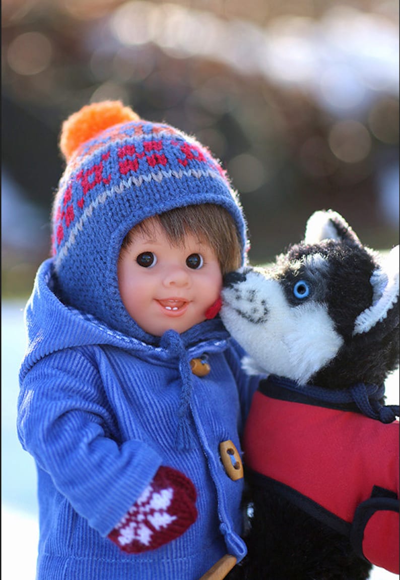 Pu&ograve; includere: Una bambola con capelli castani indossa un cappotto in velluto a coste blu, un berretto in maglia blu con motivo rosso e arancione e muffole rosse con fiocchi di neve bianchi, accanto a un cane husky di peluche bianco e nero con gilet rosso.