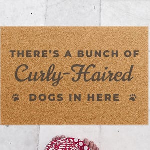 May include: A rectangular tan coir doormat featuring the dark brown text THERE'S A BUNCH OF Curly-Haired DOGS IN HERE with a small paw print icon on the left and right sides of the bottom line of text, centered on a light-colored tiled floor.