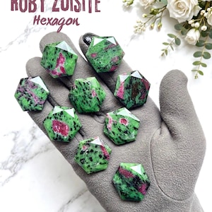 Op de afbeelding: Acht gefacetteerde zeshoekige robijn-zoisiet edelstenen met een gevlekte groene en zwarte basis met levendige magenta-roze insluitingen. De stenen zijn gerangschikt op een grijze stof. De tekst bovenaan luidt RUBY ZOISITE Hexagon.
