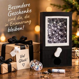 Könnte beinhalten: Ein schwarzer Kaugummiautomat gefüllt mit transparenten Kunststoffkugeln und einem weißen Drehknopf. Daneben stehen zwei braune Geschenkboxen mit schwarzen Schleifen und einem Anhänger mit der Aufschrift 'Viel Glück!'. Der Text links lautet 'Besondere Geschenke sind die, die in Erinnerung bleiben!'
