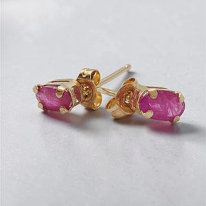 May include: A pair of stud earrings featuring oval-shaped, deep pink gemstones set in gold metal prongs. Each earring has a gold post and a butterfly-style back, showcasing a polished metallic finish and vibrant, translucent pink stones.
