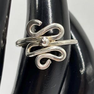 May include: A silver ring featuring a polished metal band that transitions into two symmetrical, swirling S-shaped wire designs. A small, smooth silver sphere is centered between the two loops, creating a cohesive and balanced metallic ornament.