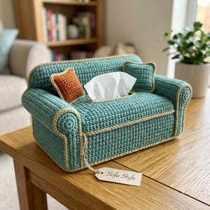 Puede incluir: Una funda de ganchillo para caja de pañuelos con forma de sofá en miniatura color verde azulado, con ribete beige, dos cojines cuadrados pequeños en verde azulado y naranja, y una etiqueta de papel con el texto Sofa Style.