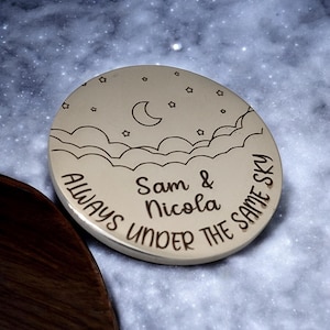 Może przedstawiać: Okrągły drewniany podkładka z wzorem półksiężyca, gwiazd i chmur. Tekst "Sam & Nicola Always Under The Same Sky" jest wygrawerowany na podkładce.