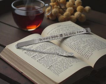 May include: An open book with a metal bookmark that reads "So many books, so little time." A cup of tea sits next to the book. Dried yellow flowers are in the background. The bookmark has a feather charm.