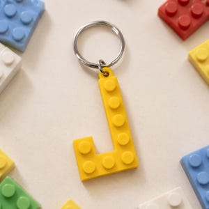 May include: A yellow letter "J" keychain made of interlocking building blocks, attached to a silver keyring. The keychain is surrounded by other colorful building blocks in blue, red, white, and yellow.