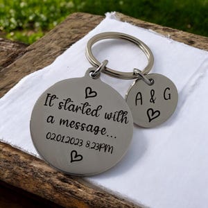 May include: A silver keychain with two round charms. The larger charm has the text "It started with a message... 02012023 8.23PM" and a heart symbol. The smaller charm has the text "A & G" and a heart symbol.