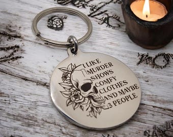 May include: Silver keychain with a skull and floral design. The text on the keychain reads "I like murder shows comfy clothes and maybe 3 people".