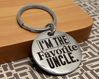 May include: A silver-colored keychain with a round pendant. The pendant has the text "I'M THE Favorite UNCLE" in black, bold font. The keychain is on a wooden surface.