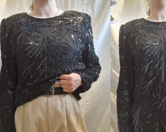 Könnte beinhalten: Ein schwarzes Langarm-Oberteil mit einem Pailletten-Blumenmuster. Das Oberteil hat einen runden Ausschnitt und lange Ärmel. Das Oberteil wird mit einer beigefarbenen Hose getragen.