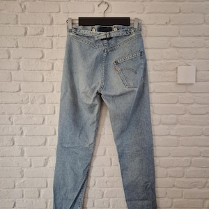 Könnte beinhalten: Hellblaue Jeans mit hoher Taille und Gürtel. Die Jeans hängen an einem Kleiderbügel vor einer weißen Backsteinmauer. Die Gesäßtasche hat ein rotes Etikett.