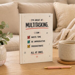 Op de afbeelding: Een crèmekleurig hardcover boek met zwarte tekst: I'M GREAT AT MULTITASKING. I CAN: WASTE TIME, BE UNPRODUCTIVE, PROCRASTINATE, ALL AT ONCE. De tekst bevat rode en blauwgroene vinkjes, een rood hartje en kleine sterillustraties.
