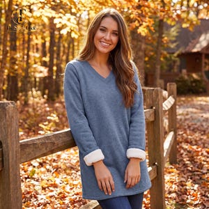 May include: A long-sleeved, heather blue tunic top featuring a V-neckline and white faux-shearling cuffs. The garment has a straight hemline and a soft, fleece-like texture, providing a layered appearance at the wrists with the white lining.