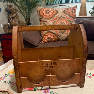 May include: A vintage wooden caddy with a top handle and two circular cutouts at the base. The front panel features the black printed text ESQUIRE FOOTMAN De Luxe PAT PEND. The structure is made of light brown wood with visible grain and wear.