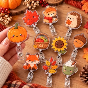 May include: A collection of nine acrylic badge reels featuring autumn-themed designs including a smiling pumpkin, a red maple leaf, a bear with a coffee cup, a hedgehog, a basket of apples, a sunflower, a fox, a branch of leaves, and a mug.