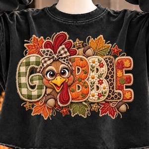 Op de afbeelding: Een zwart cropped sweatshirt met een grote afbeelding die GOBBLE spelt. De letter O is vervangen door een cartoonkalkoen met een bril en een strik. De letters bevatten patronen van groen ruitjesmotief, oranje pompoenen, witte madeliefjes en luipaardprint.