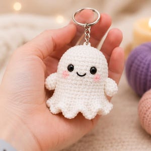May include: A white crocheted ghost keychain featuring black plastic eyes, a stitched black mouth, and small pink embroidered cheeks. The ghost is attached to a silver-toned metal split ring by a short chain, creating a small, soft, and textured accessory.