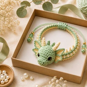 May include: A crochet bracelet featuring a light green dragon head with cream-colored horns and ears. The band is made of light green and cream yarn, accented with small white pearls and green beads, finished with an adjustable sliding knot closure.