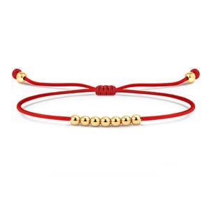 May include: Red string bracelet featuring six gold-colored beads. The bracelet has an adjustable sliding knot closure and gold-colored accents at the ends. This minimalist bracelet is a simple and elegant accessory.