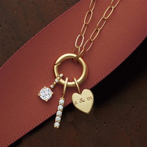 Puede incluir: Un collar de oro con un c&iacute;rculo de encanto, un encanto de coraz&oacute;n con la inscripci&oacute;n "A & M", y dos encantos de diamantes. El collar est&aacute; sobre una cinta roja.