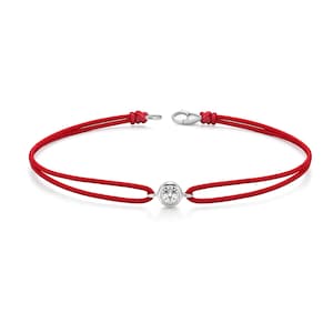 May include: A red cord bracelet with a single, round, clear gemstone in the center. The bracelet has a silver clasp and is displayed on a white background.