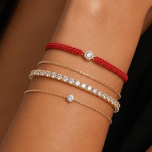 May include: A set of four bracelets consisting of one red woven cord bracelet with a central clear stone, two thin gold-tone chain bracelets, and one gold-tone tennis bracelet featuring a continuous row of sparkling clear round-cut stones.