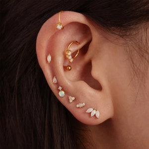 May include: A collection of gold-toned earrings with white opal stones displayed on an ear. The earrings include studs, hoops, and a helix earring. The opal stones have a pearlescent sheen.