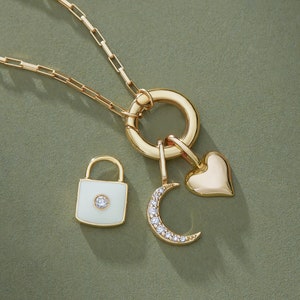 Puede incluir: Un collar de oro con un c&iacute;rculo de encanto, un encanto de coraz&oacute;n, un encanto de luna creciente y un encanto de candado con una cara de esmalte blanco y una peque&ntilde;a piedra transparente.