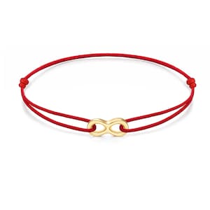 May include: A red cord bracelet featuring a central gold-toned infinity symbol charm. The bracelet is constructed from a thin, smooth red string that loops through the metal infinity charm, with adjustable knots on each side of the central piece.