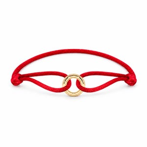 May include: A red cord bracelet featuring a polished gold-tone circular ring at the center. The cord is looped through the ring and secured with two knots on either side, creating a continuous band design with a smooth, metallic finish on the ring.