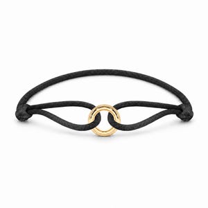 May include: A black cord bracelet featuring a single polished gold-tone circular ring at the center. The cord is looped through the ring and secured with knots on either side, creating a simple, continuous design with a smooth, metallic finish.