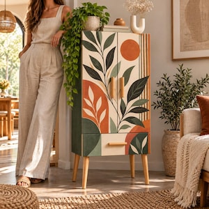 Puede incluir: Un armario de madera alto y rectangular con dos puertas y un cajón inferior, decorado con un patrón botánico pintado de hojas verde oscuro, arcos de terracota y un sol naranja circular sobre un fondo crema con tiradores de madera clara.