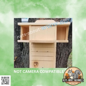 May include: A light-colored wooden squirrel feeder with a central compartment and two side feeding areas. The front features a branded logo with the text NAS and an acorn icon. The text NOT CAMERA COMPATIBLE is printed below.