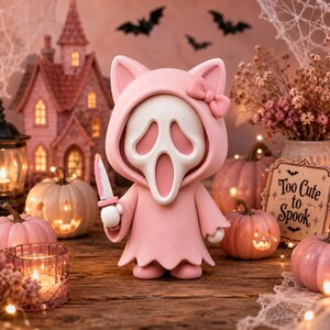 May include: A pink figurine wearing a hooded cloak with cat ears and a bow, featuring a white ghost mask with a long open mouth. The figure holds a small knife with a pink handle. The design is smooth and matte, standing upright on a wooden surface.