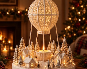 May include: A decorative tabletop display featuring a central hot air balloon structure with an intricate lattice design, suspended above a basket holding a lit candle, surrounded by miniature snow-dusted houses and evergreen trees on a round base.