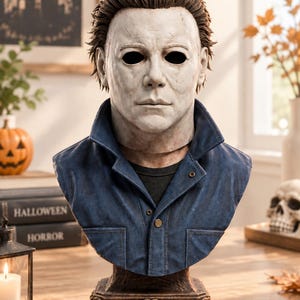 Peut inclure: Un buste représentant un personnage portant un masque blanc vieilli aux orbites sombres et des cheveux bruns. La figurine porte une veste bleue à col sur une chemise noire, fixée sur un socle décoratif marron avec une plaque nominative vide.